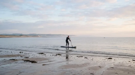 Un débutant en combinaison néoprène rame en paddle sur une baie normande calme au lever du jour, avec des plages de sable humide à marée basse au premier plan.