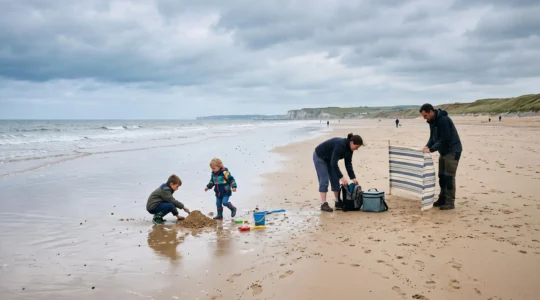 Une famille avec deux enfants joue sur une plage normande sous un ciel couvert, vêtue de couches superposées et de bottes, avec un pare-vent planté dans le sable.