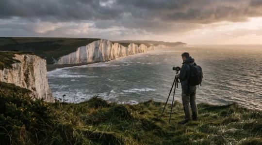 Un photographe avec un trépied observe une côte normande à falaises sous un ciel changeant, à la lumière rasante de fin de journée.